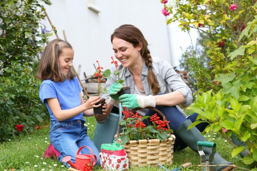 An adult woman and a young girl are gardening together in a lush, well-maintained outdoor garden space. The woman, wearing a grey shirt, jeans, and green gardening gloves, is smiling as she holds a small potted plant with red flowers. The girl, dressed in a blue t-shirt and denim shorts, is sitting on the grass, using her hands to handle the plant, and appears engaged and happy. Around them, there are various garden elements including a wicker basket, a red watering can, and a decorative fabric-covered container. The garden features a neatly trimmed lawn, tall green bushes, flowering plants with pink and red blossoms, and a backdrop of a white wall or fence partially obscured by foliage. The scene is well-lit with natural daylight, suggesting a bright, pleasant day, typical of outdoor gardening activities in Fulham, London. The image naturally aligns with gardening services offered by Hedge Trimming Fulham, emphasizing the nurturing and recreational aspects of outdoor garden maintenance in a peaceful residential setting.