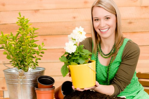 A woman with shoulder-length blonde hair and a bright smile sits outdoors in a garden setting, holding a yellow flowerpot containing a lush green plant with white flowers. She is dressed in a green top with a brown cardigan and wearing green beaded necklaces. In front of her on a wooden table are various gardening items: a metallic pot with a small, healthy green shrub, empty terracotta pots, and a black container. The background features a wooden fence and a well-maintained lawn with grass of medium density and vibrant green colour. The garden environment appears well-kept, with a mix of potted plants and natural elements, reflecting a pleasant outdoor space suitable for gardening services in Fulham or nearby areas. The weather looks clear with natural daylight illuminating the scene, emphasizing the freshness of the plants and the tidy, inviting outdoor area.